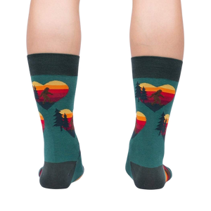 A pair of green socks with a pattern of red, orange, and yellow hearts. The hearts have a silhouette of a sasquatch inside them, along with pine trees and a mountain range.