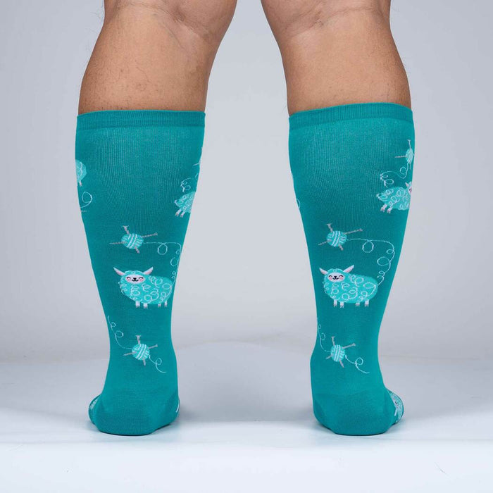 A pair of teal knee-high socks with a pattern of white sheep and blue yarn balls.