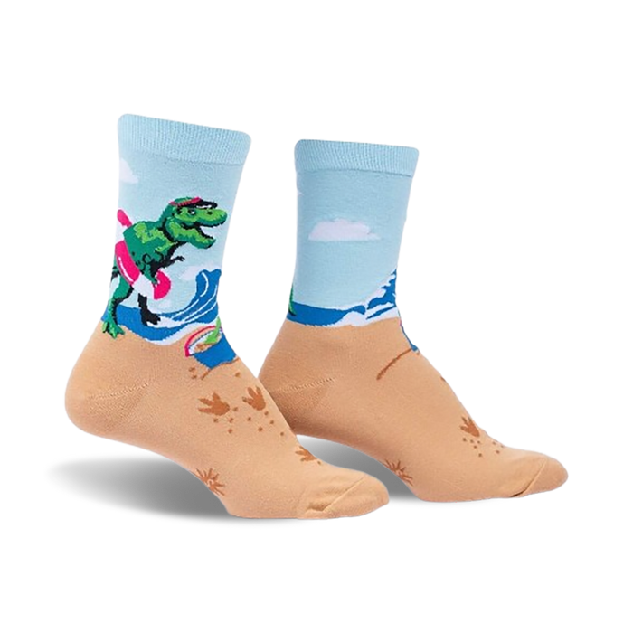 A pair of crew socks showcasing a T-Rex surfing on a beach.  The socks are light blue and tan with green, pink, and white accents.