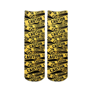 Pair of crew socks with a repeating caution tape pattern in black and yellow.  The design features the word 'CAUTION' printed on the tape.