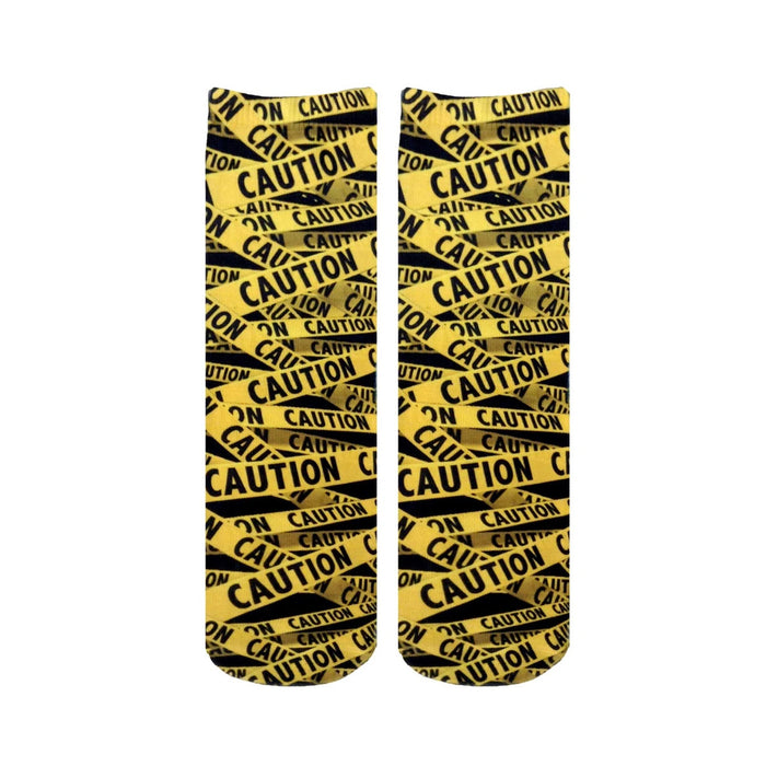 Pair of crew socks with a repeating caution tape pattern in black and yellow.  The design features the word 'CAUTION' printed on the tape.