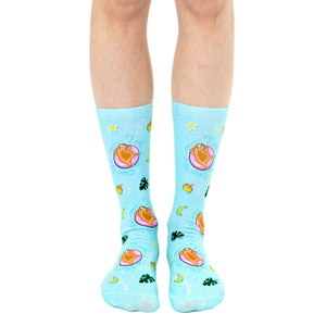 A pair of light blue crew socks with a fun design. Sloths lounging on pink inner tubes in a pool are repeated against a tropical background with yellow stars, moons, and leaves.