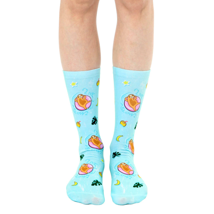 A pair of light blue crew socks with a fun design. Sloths lounging on pink inner tubes in a pool are repeated against a tropical background with yellow stars, moons, and leaves.