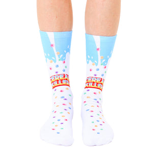A pair of white crew socks with a light blue milk splash design and colorful cereal pieces. The text 'Cereal Killer' is prominently displayed.