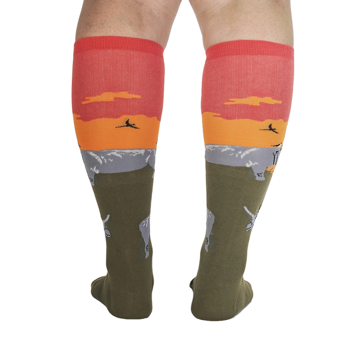 A pair of green knee-high socks with an orange and yellow sunset scene with silhouettes of large animals at the bottom.