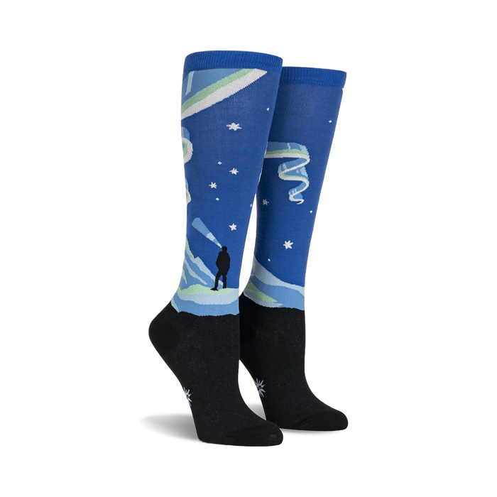 Pair of knee-high socks showcasing a Northern Lights design. Blue, green, and black colors are featured, along with a silhouette of a person watching the lights.