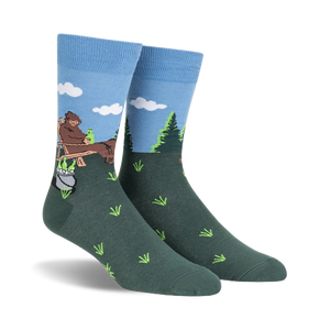 Crew socks in blue and green, depicting a cartoon Bigfoot relaxing in a wooden chair outdoors with a pot and bottles beside him.