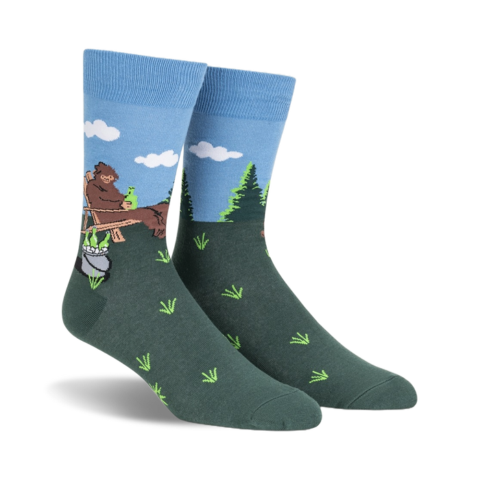Crew socks in blue and green, depicting a cartoon Bigfoot relaxing in a wooden chair outdoors with a pot and bottles beside him.