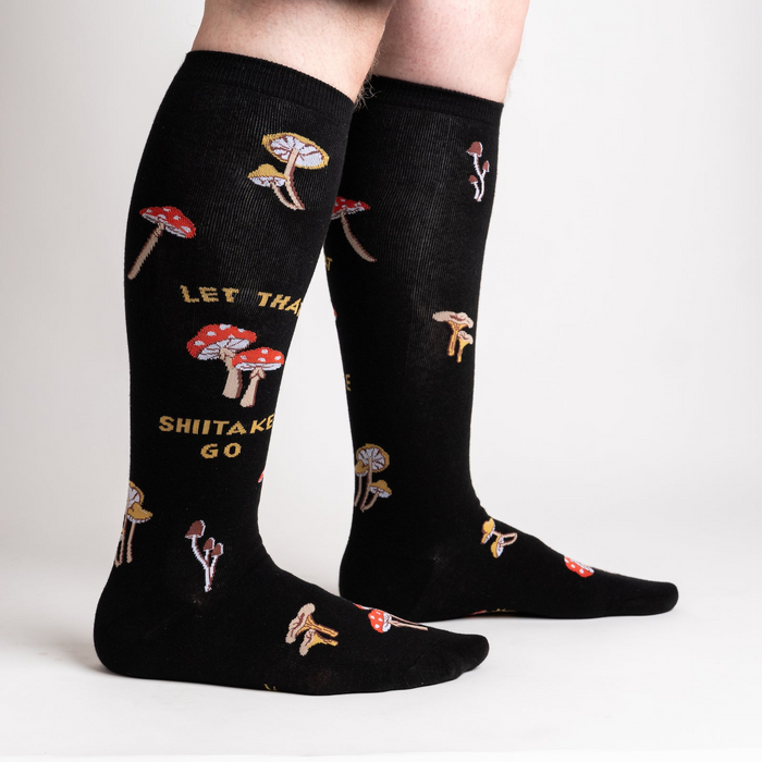 Lifestyle shot showing the 'Let That Shiitake Go' socks on a person's legs. The socks are black with a mushroom and text pattern.