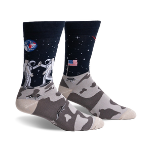 Pair of crew socks; navy blue with astronauts high-fiving on the moon, a US flag, and a lunar landscape. The foot features a grey moon rock pattern.