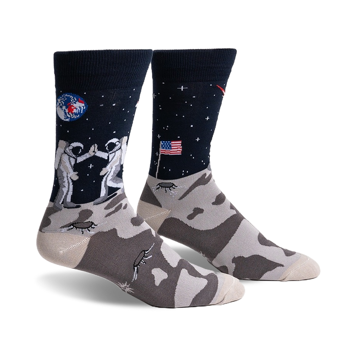Pair of crew socks; navy blue with astronauts high-fiving on the moon, a US flag, and a lunar landscape. The foot features a grey moon rock pattern.