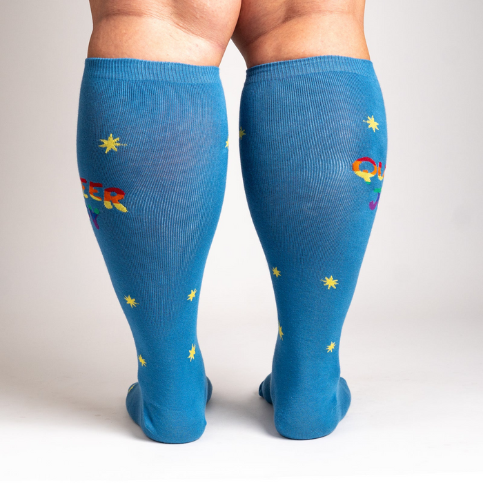 Rear view of model wearing blue knee-high socks featuring 'Queer Joy' in rainbow and yellow stars.