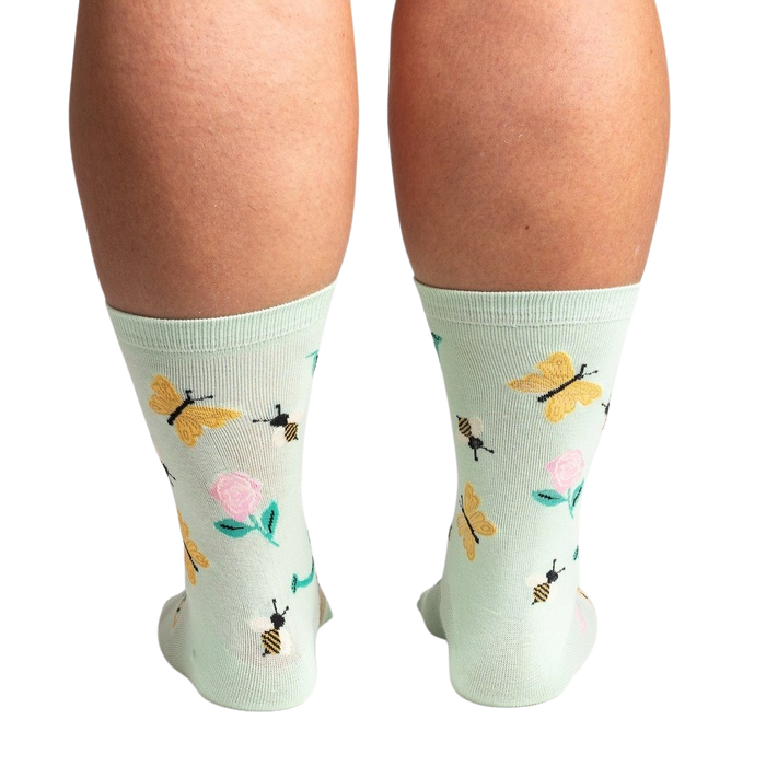 A pair of light green socks with a pattern of pink roses, yellow butterflies, and black bees.