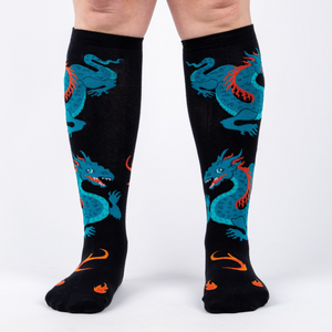 Front view of model wearing a pair of black knee-high socks with teal dragons and orange flames.