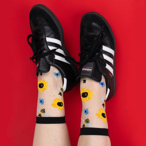Lifestyle shot: Sheer socks with sunflower and bee design worn with black Adidas sneakers against a red background.