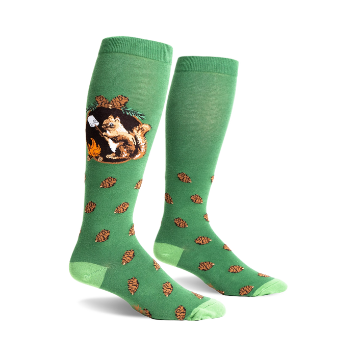 A pair of green knee-high socks with a whimsical design. One sock shows a squirrel roasting a marshmallow by a campfire; the other has scattered pine cones.