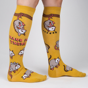 Lifestyle shot: Model wearing a pair of mustard yellow knee-high socks with possum designs.