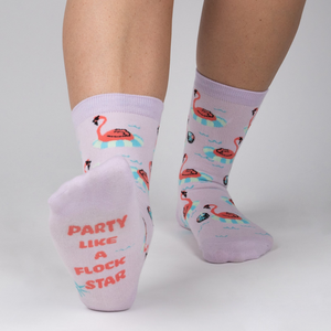 Close-up of a person's feet showcasing the 'Party Like A Flock Star' text on the sole of a lavender crew sock, accompanied by a flamingo design.