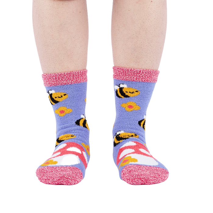 Person wearing fuzzy periwinkle slipper socks with bee, mushroom, and flower patterns, front view.