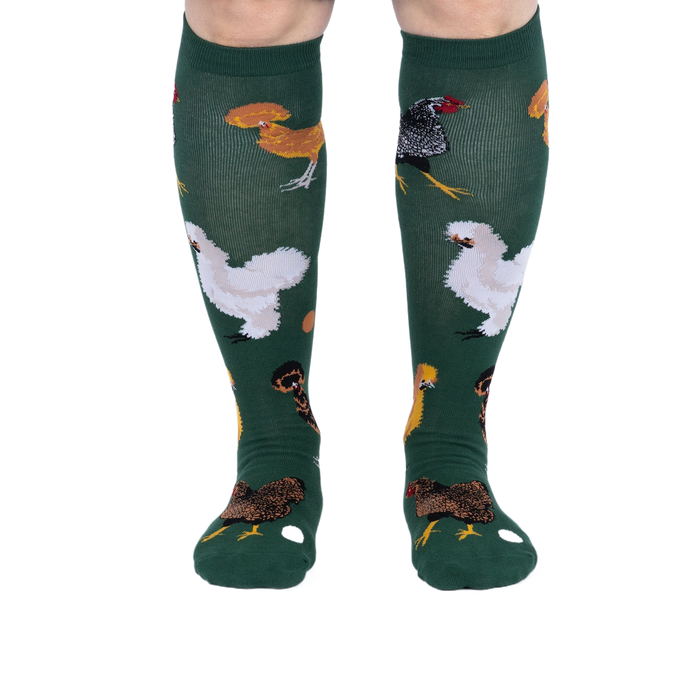 Person wearing dark green knee-high socks patterned with various chickens and eggs, standing, front view.