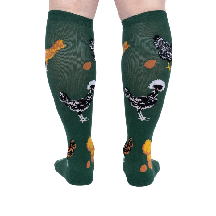 Person wearing dark green knee-high socks patterned with various chickens and eggs, standing, back view.
