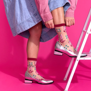 Person in sheer pink cowboy boot socks, silver flats, denim skirt, and pink shirt, standing on a step stool against a bright pink wall.