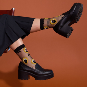 Person wearing sheer black cat socks and black chunky loafers, seated with feet up against an orange background.