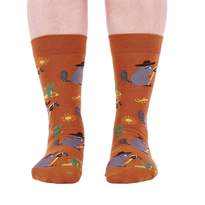 Front view of a person wearing burnt orange crew socks with a repeating pattern of raccoon cowboys, cacti, suns, and lizards.