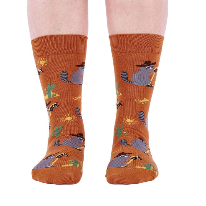 Front view of a person wearing burnt orange crew socks with a repeating pattern of raccoon cowboys, cacti, suns, and lizards.