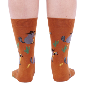 Back view of a person wearing burnt orange crew socks with a repeating pattern of raccoon cowboys, cacti, suns, and lizards.