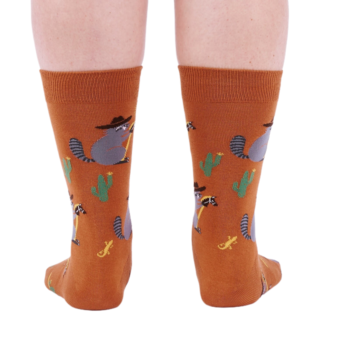Back view of a person wearing burnt orange crew socks with a repeating pattern of raccoon cowboys, cacti, suns, and lizards.