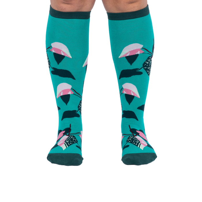 Front view of a person's legs wearing teal knee-high socks with pink and dark green philodendron leaves.