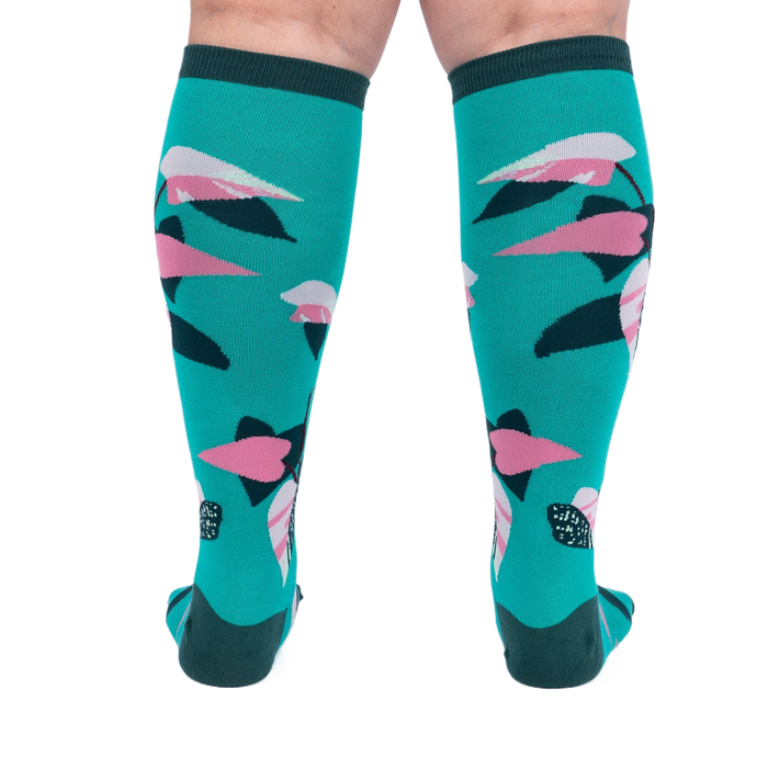 Back view of a person's legs wearing teal knee-high socks with pink and dark green philodendron leaves.