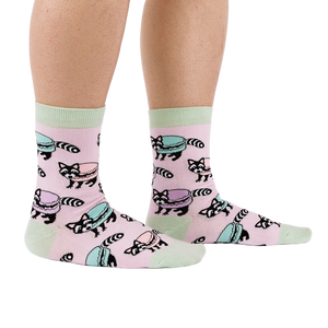 Side view of a person wearing pink crew socks with a pattern of raccoons and pastel macarons, showing light green cuffs and heels.