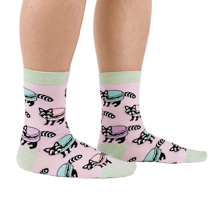 Side view of a person wearing pink crew socks with a pattern of raccoons and pastel macarons, showing light green cuffs and heels.