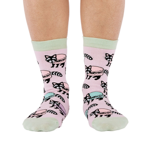 Front view of a person wearing pink crew socks adorned with a pattern of cartoon raccoons and light blue and purple macarons.