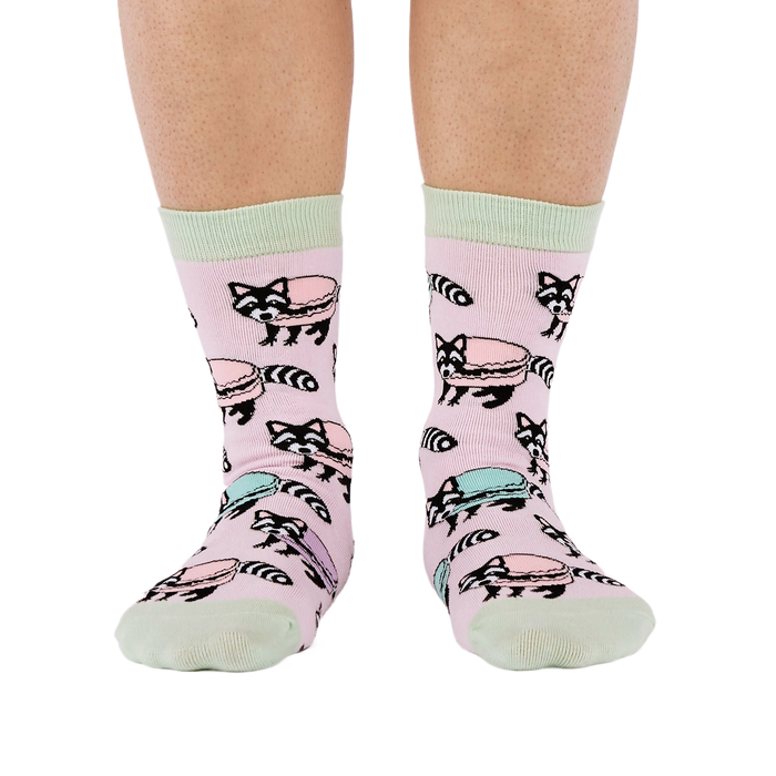 Front view of a person wearing pink crew socks adorned with a pattern of cartoon raccoons and light blue and purple macarons.