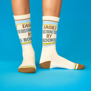Person's legs wearing cream crew socks with 'Easily Distracted By Books' text, standing on a blue surface.