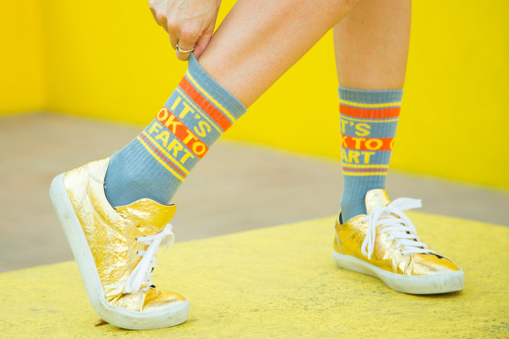 Lifestyle shot: Person wearing 'It's Ok to Fart' socks with gold shoes against a yellow background.