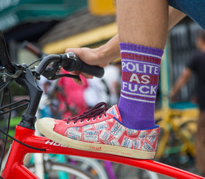Lifestyle photo: Person wearing one purple 'Polite As Fuck' sock with red shoes and riding a bicycle.