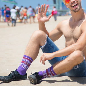 Lifestyle photo: Man sitting on the beach wearing the purple 'Polite As Fuck' socks and ripped denim shorts.