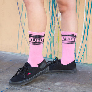 Lifestyle shot of a person wearing the pink 'Butts' socks with black shoes against a colorful wall.