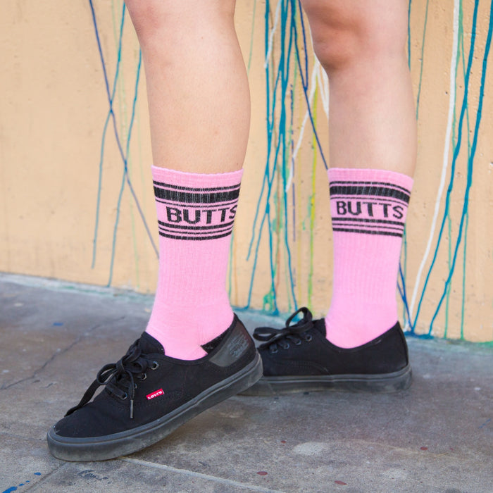 Lifestyle shot of a person wearing the pink 'Butts' socks with black shoes against a colorful wall.