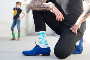 A person kneeling, showcasing the socks and blue shoes; another person is blurred in the background.