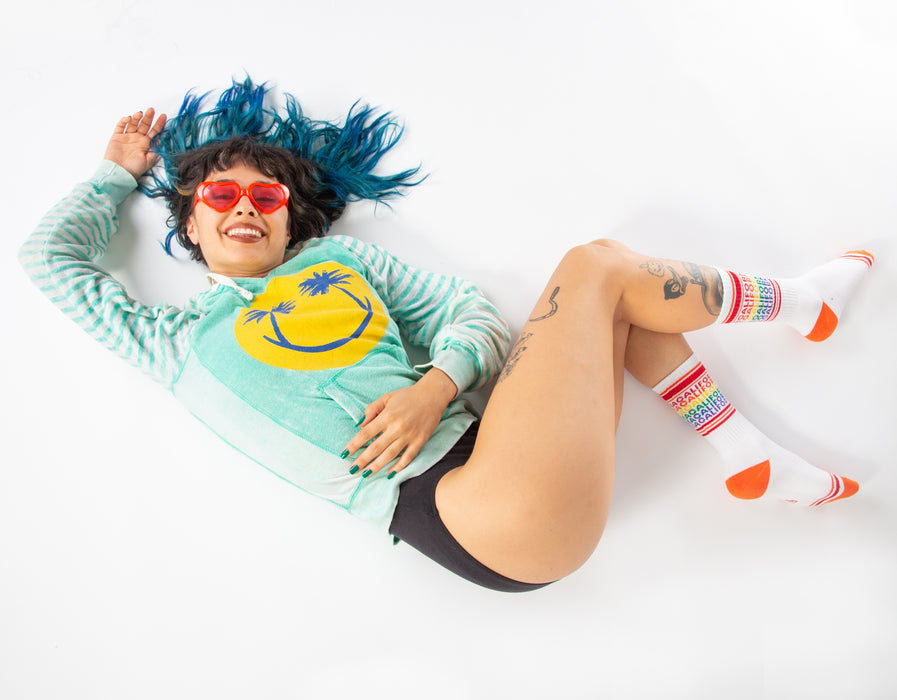 A young woman is lying on her back against a white background. She has blue hair and is wearing red heart-shaped sunglasses, a green sweatshirt, black underwear, and multi-colored socks. She has tattoos on her legs and is smiling at the camera.