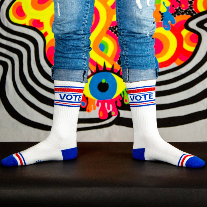 A pair of white socks with red and blue stripes at the top and blue heels and toes. The word VOTE is knit into the front of the socks in red, white, and blue.