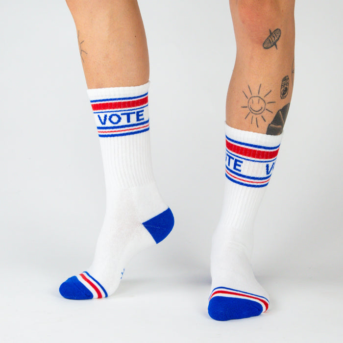 A pair of white socks with red and blue stripes at the top and blue heels and toes. The word VOTE is knit into the front of the socks in red, white, and blue.