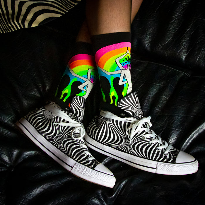A pair of zebra-striped high-top Converse sneakers with rainbow laces are being worn with a pair of colorful socks that have a dancing skeleton on them. The person is sitting on a black leather couch with a zebra-striped pattern.