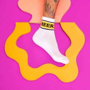 A pair of white socks with the word BEER in yellow letters on the leg portion. The socks are being worn by a person who is standing on a black and white checkered floor.