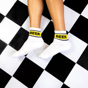 Flatlay showing two white socks with yellow 'BEER' bands on a black and white checkered floor.